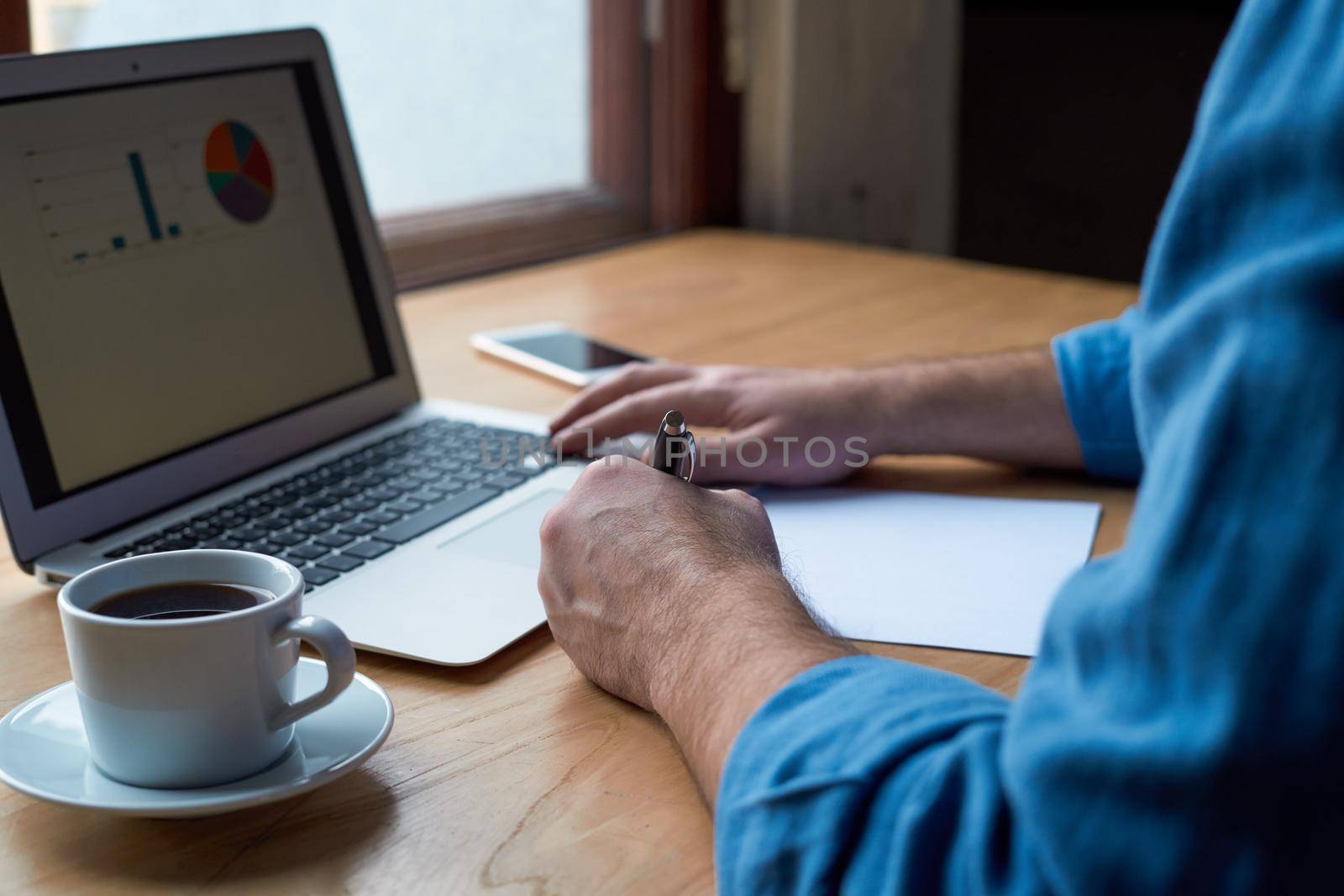 Unrecognizable man writes plan on paper and looks at charts on computer screen, laptop. A man with casual clothes in blue shirt sits in office in front of window, cup of coffee