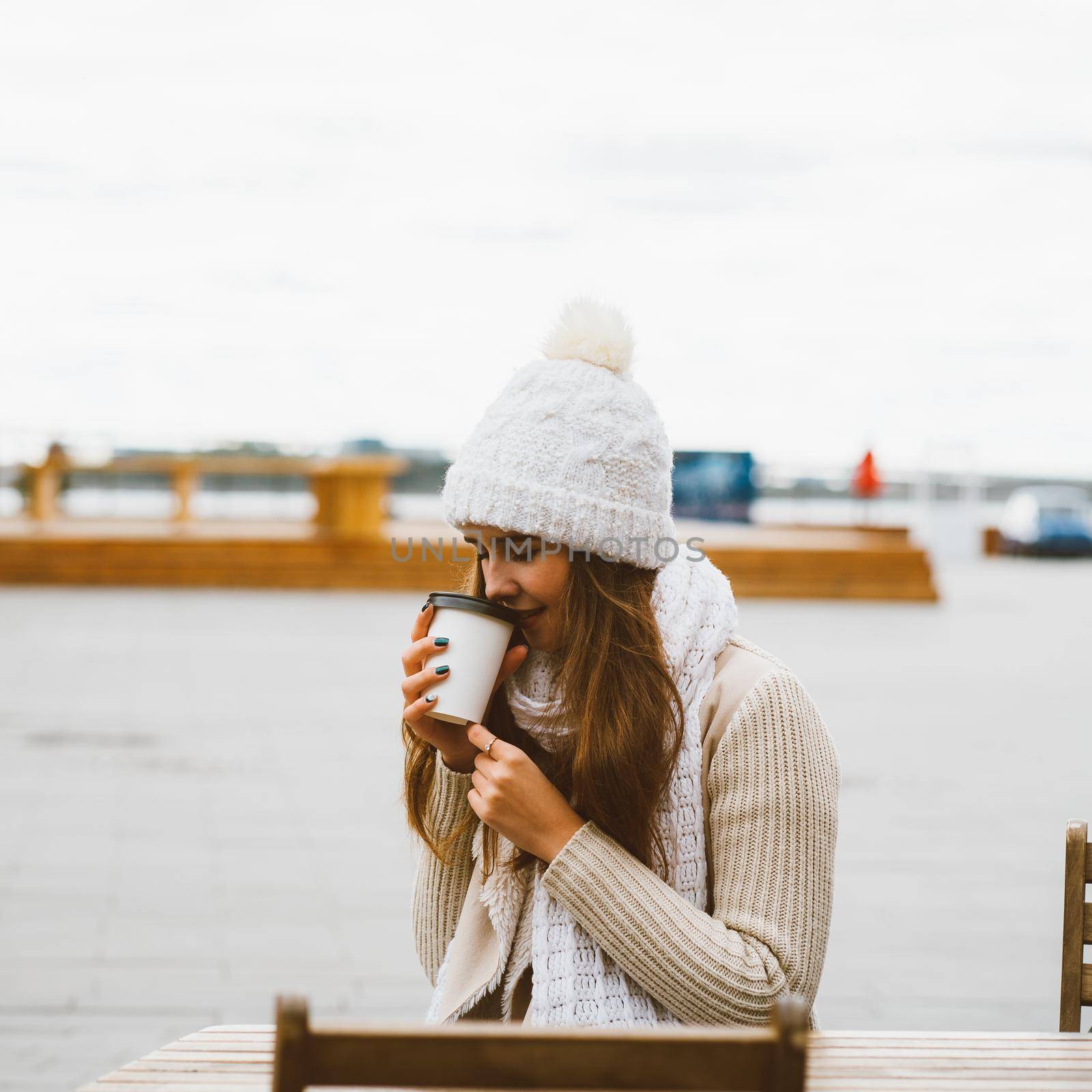 Beautiful young girl drinking coffee, tea from plastic mug in a autumn, winter. A woman with long hair in warm clothes sitting on waterfront on Baltic sea in port, warmed by hot drink