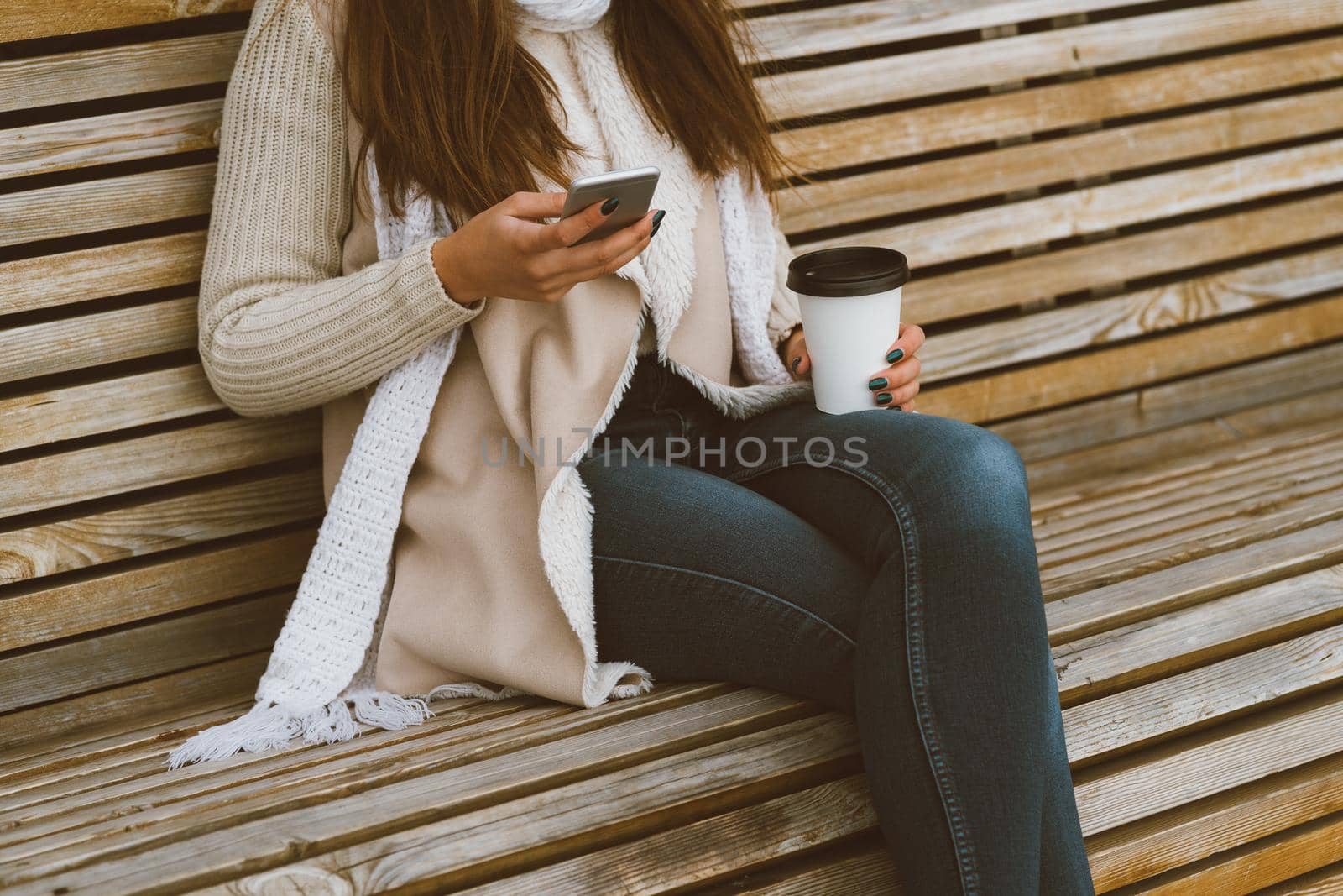 Unrecognizable beautiful young girl drinking coffee, tea from a plastic mug in autumn, winter and talking on mobile phone. Woman with long hair sitting on bench in autumn or winter, basking in hot drink, copy space