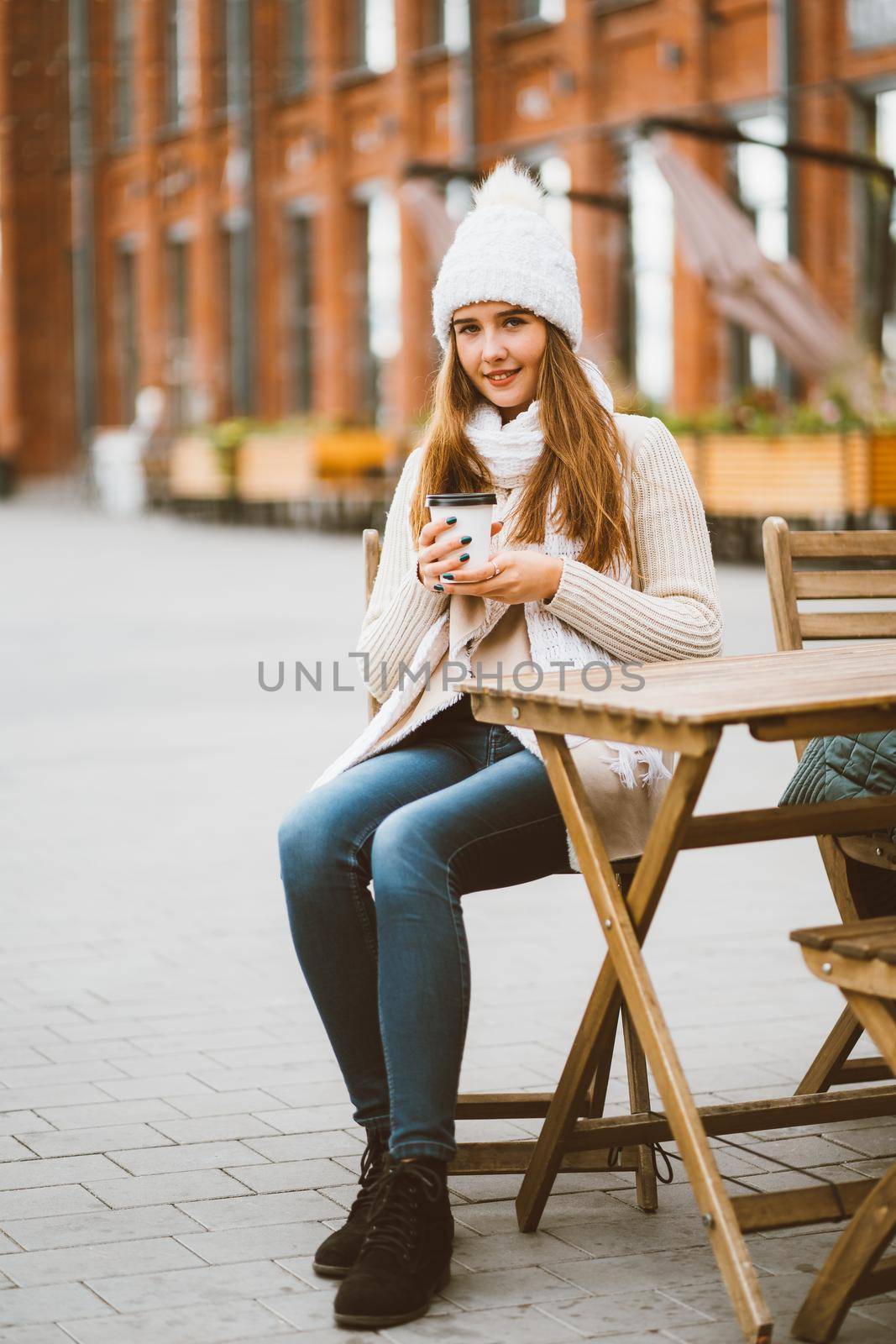 Beautiful young girl drinking coffee, tea from plastic reusable mug in autumn, winter. Woman with long hair, in warm clothes sitting in street cafe, warmed by hot drink, vertical by NataBene