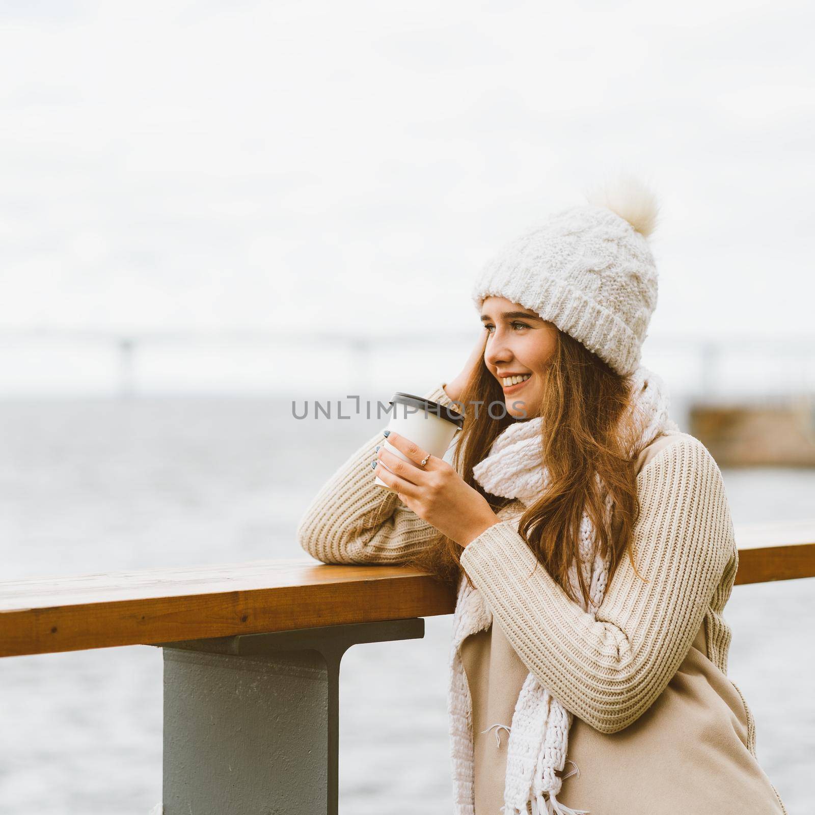 Beautiful young girl drinking coffee, tea from plastic mug in autumn, winter. A woman with long hair stands on waterfront on Baltic sea in port and waiting for ferry, heated by a hot drink, copy space