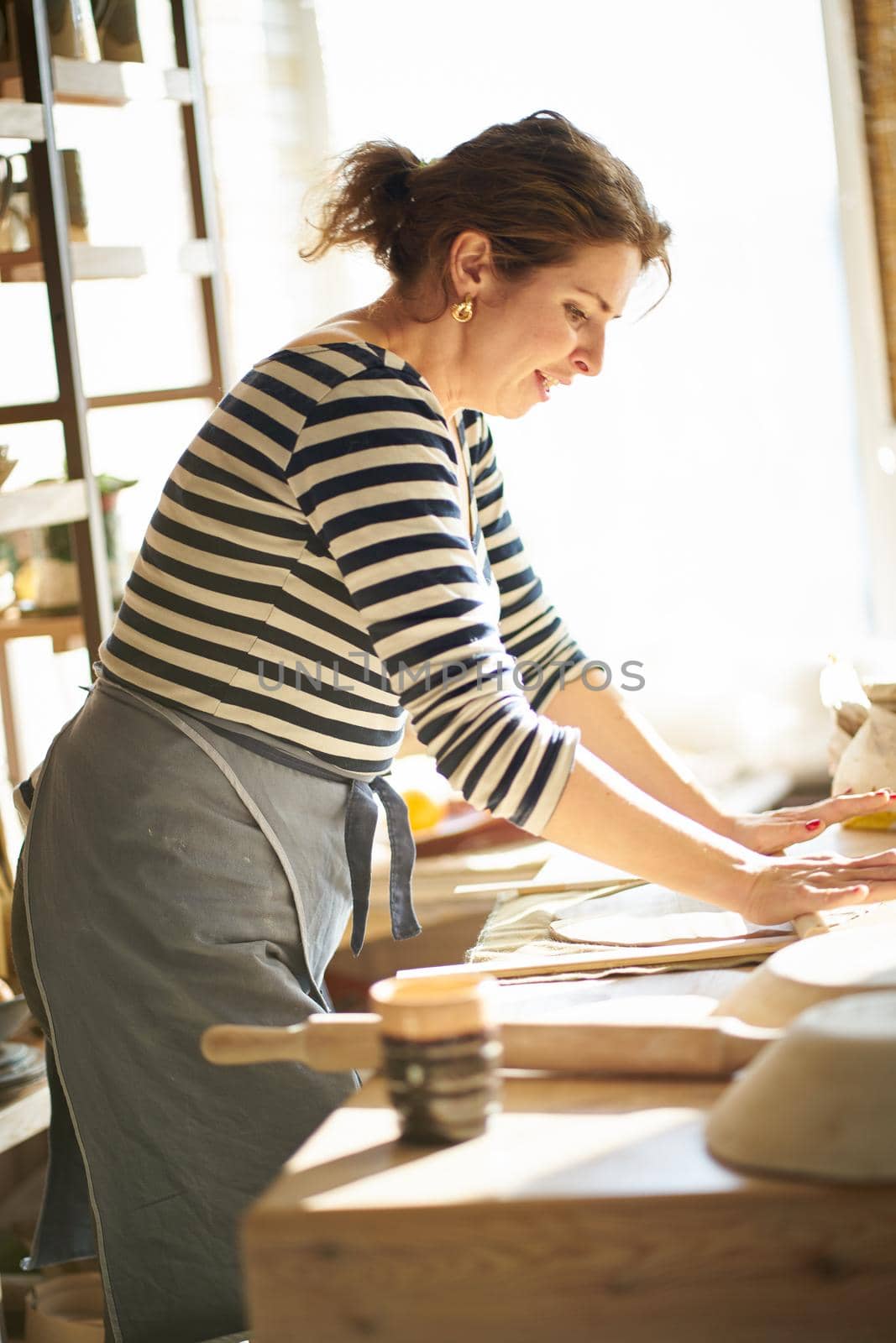 Beautiful happy woman making ceramic ware in workplace, laughing, smiling in sun light. Concept for woman in freelance, business, hobby