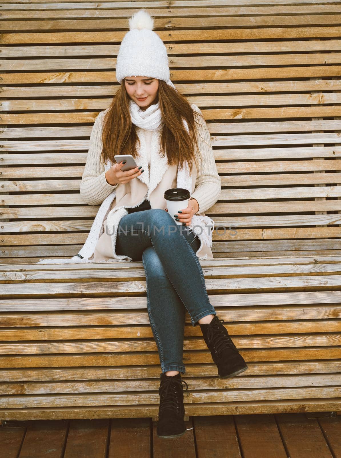 Beautiful young girl drinking coffee, tea from a plastic mug in autumn, winter and talking on a mobile phone. Woman with long hair sitting on a bench in autumn or winter, basking in a hot drink, copy space, vertical