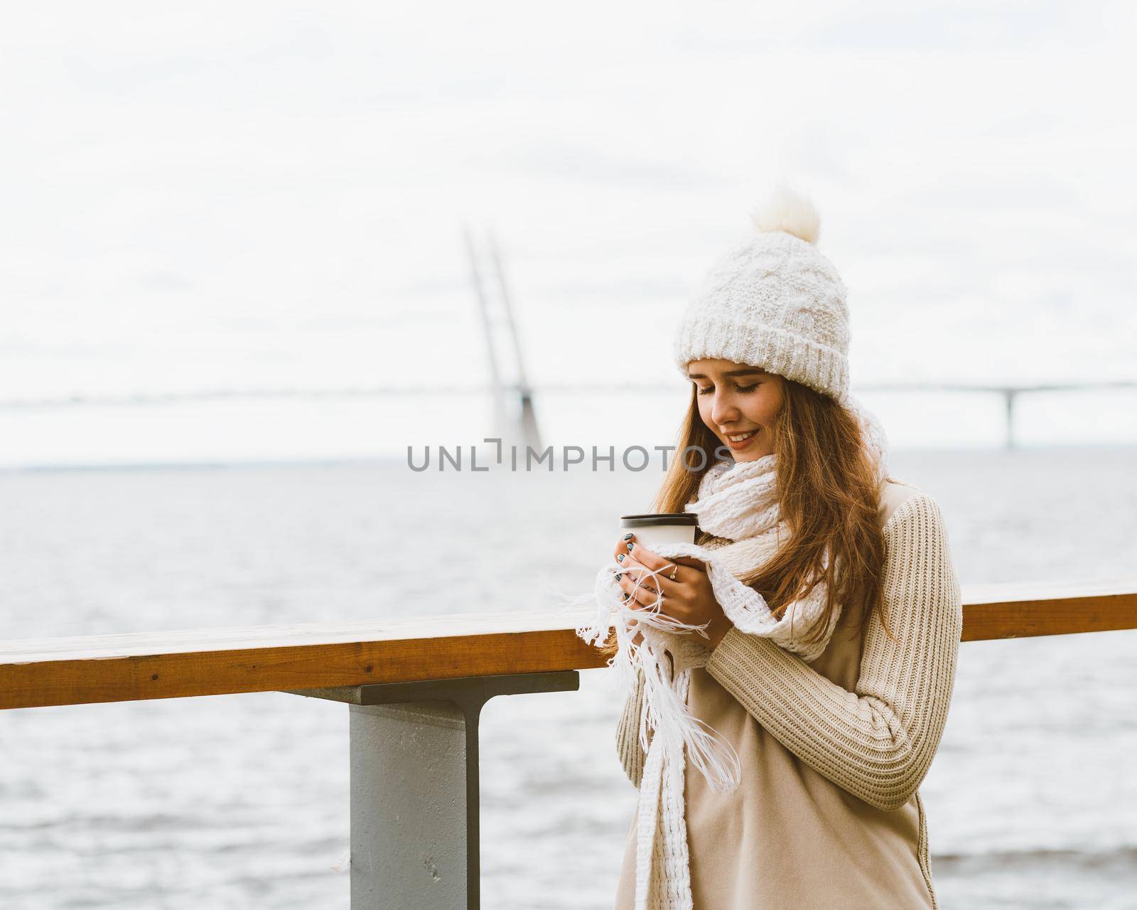 Beautiful young girl drinking coffee, tea from plastic mug in autumn, winter. A woman with long hair stands on waterfront on Baltic sea in port and waiting for ferry, heated by a hot drink, copy space