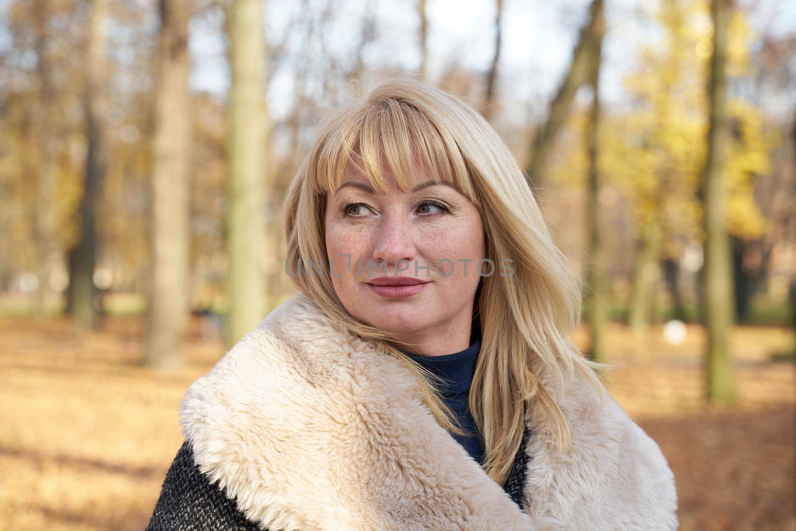 Happy blonde mature woman in autumn park and thinking. Beautiful woman is relaxing in nature on a sunny day. Close up portrait of middle aged woman smiling and daydreaming.