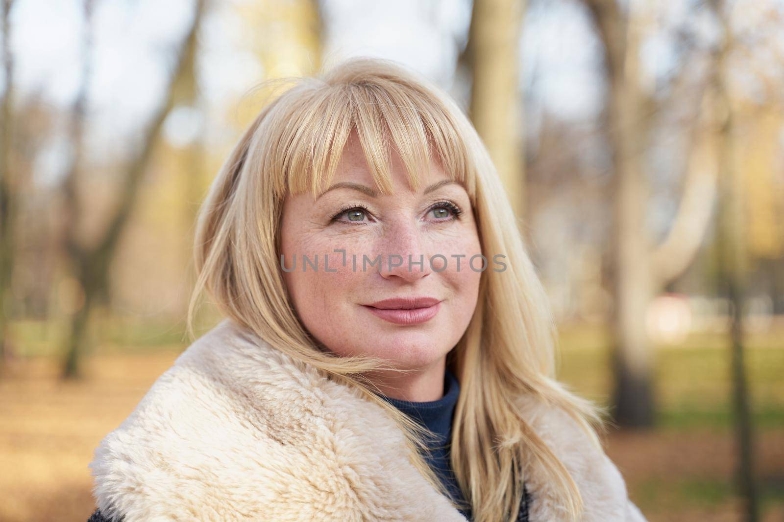 Closeup portrait of happy blonde mature woman is standing in autumn the park and thinking. Beautiful woman is relaxing in nature on sunny day. Portrait of middle aged woman smiling and daydreaming.