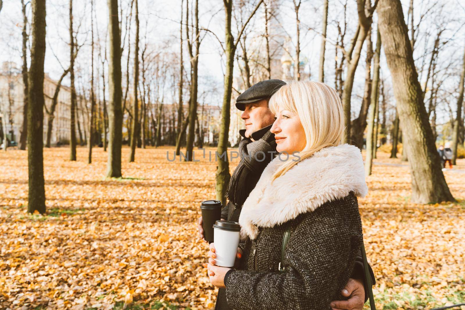 Happy blonde mature woman and handsome middle-aged brunette man walk in park and drink coffee from reusable cup. A loving couple of 45-50 years old walks in autumn park in warm clothes, in coat