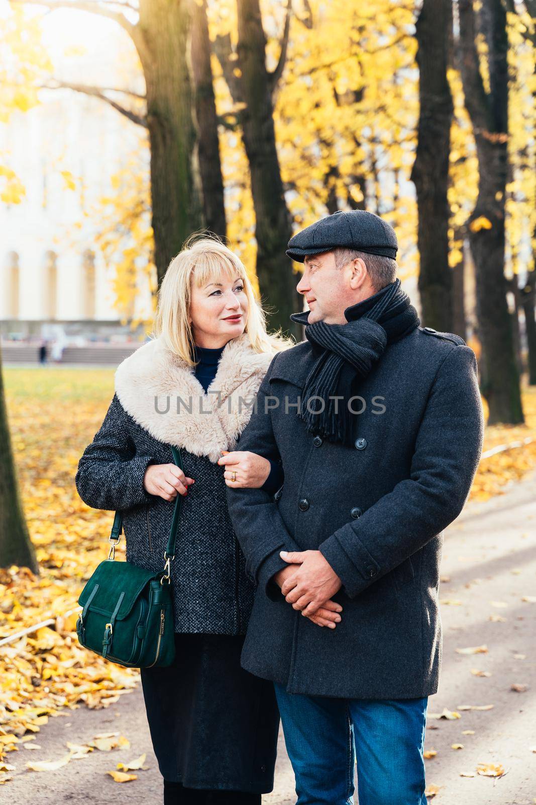 Happy blonde mature woman and handsome middle-aged brunette man walk in park, looking at each other. A loving couple of 45-50 years old walks in the autumn park in warm clothes