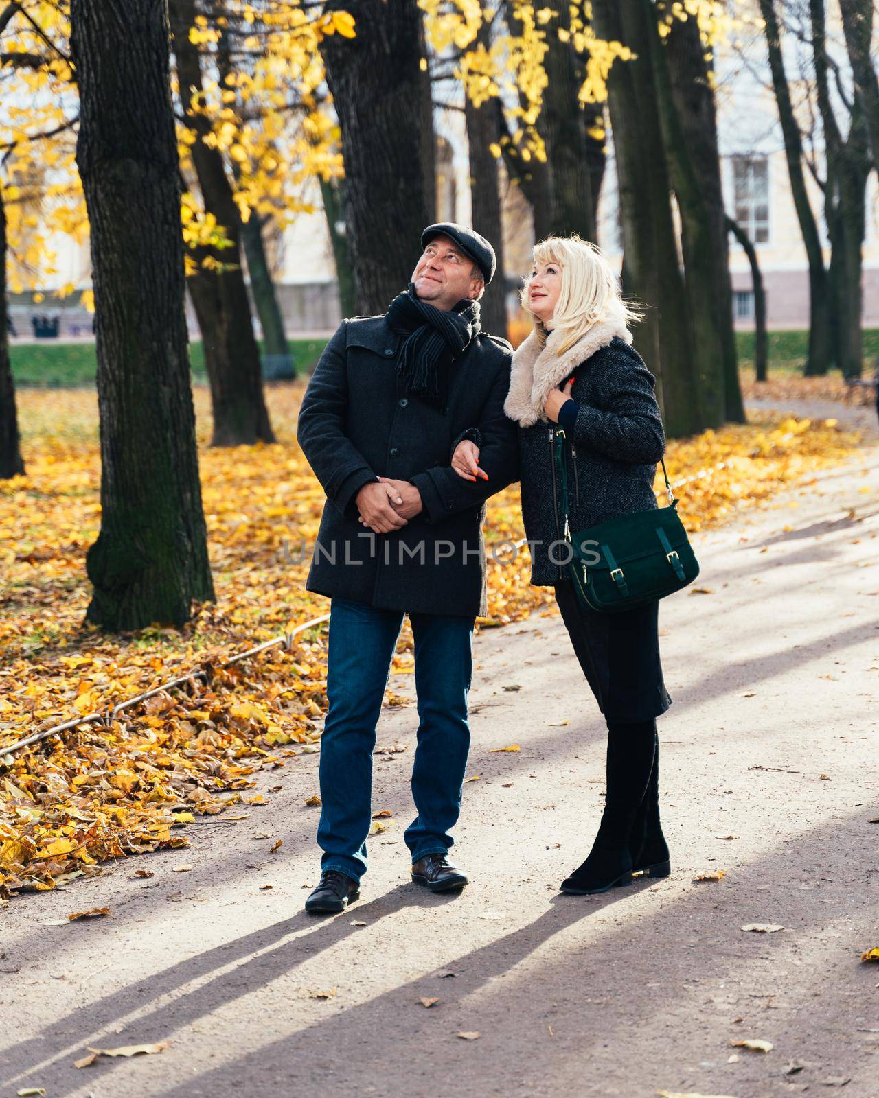 Happy blonde mature woman and beautiful middle-aged brunette, look up to sky, walking in park. Loving couple of 45-50 years old walks in the autumn park in warm clothes