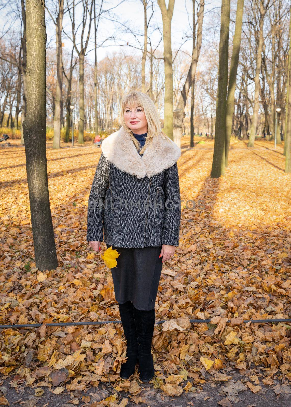 Happy blonde mature woman is standing in autumn park and thinking. Beautiful woman is relaxing in nature on a sunny day. Portrait of a middle aged woman smiling and daydreaming.