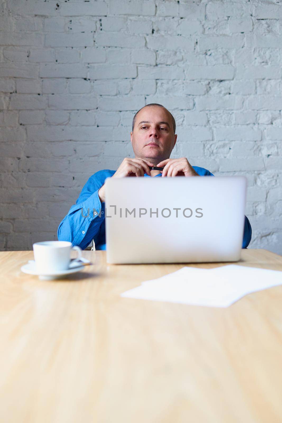 Handsome handsome mature man sitting at table in front of laptop and listening to a subordinate. Man with casual clothes in blue shirt in the office in front of window, vertical, copy space