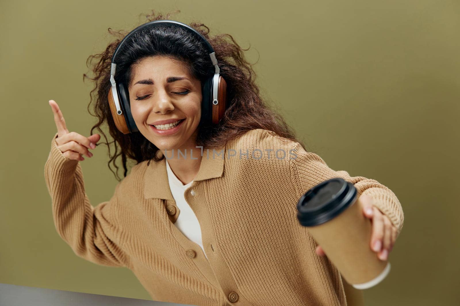 COOL MOOD CONCEPT. Enjoyed tanned curly Latin female in headphones with coffee cup to-go dance sit at table isolated over olive green background close eyes. Copy space Mockup Banner. Fashion offer