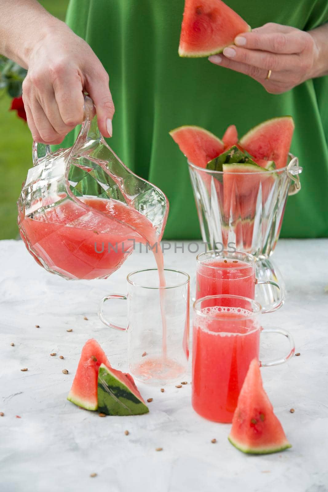 a woman in a green dress pours a red refreshing drink from the pulp of a watermelon into a glass glass, bright natural background
