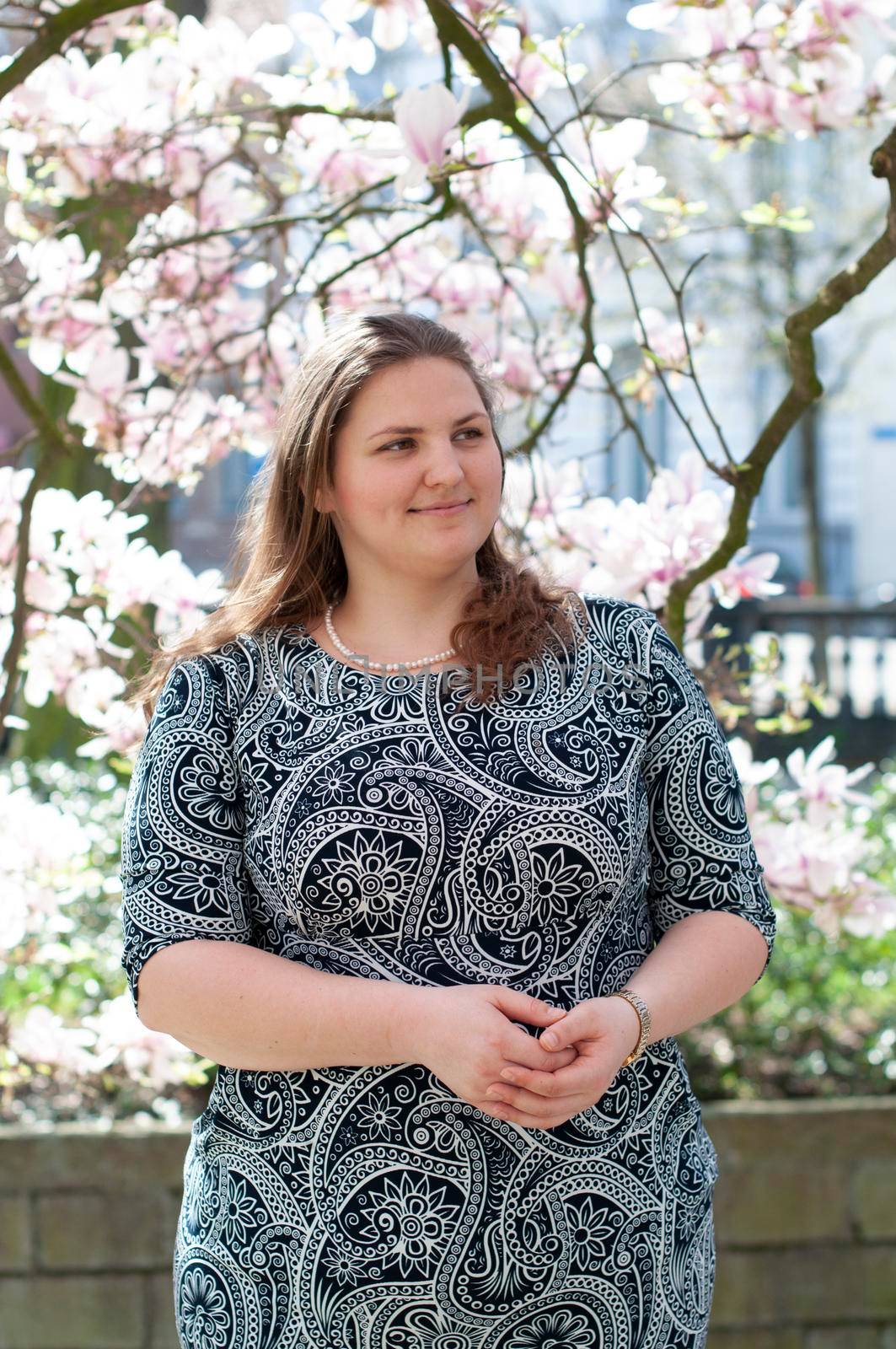 A plump, confident woman stands under a blooming magnolia and smiles. young millennial woman with brown curly hair smiling and waiting for a friend for a date. A full girl enjoys flowering and spring.