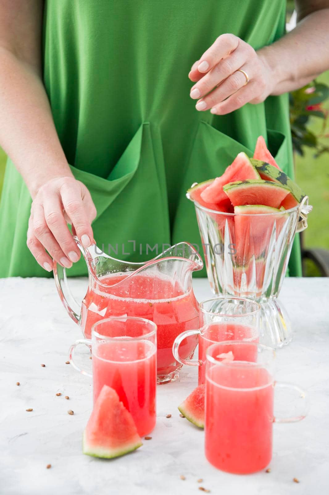 a woman in a green dress pours a red refreshing drink from the pulp of a watermelon into a glass glass, bright natural background