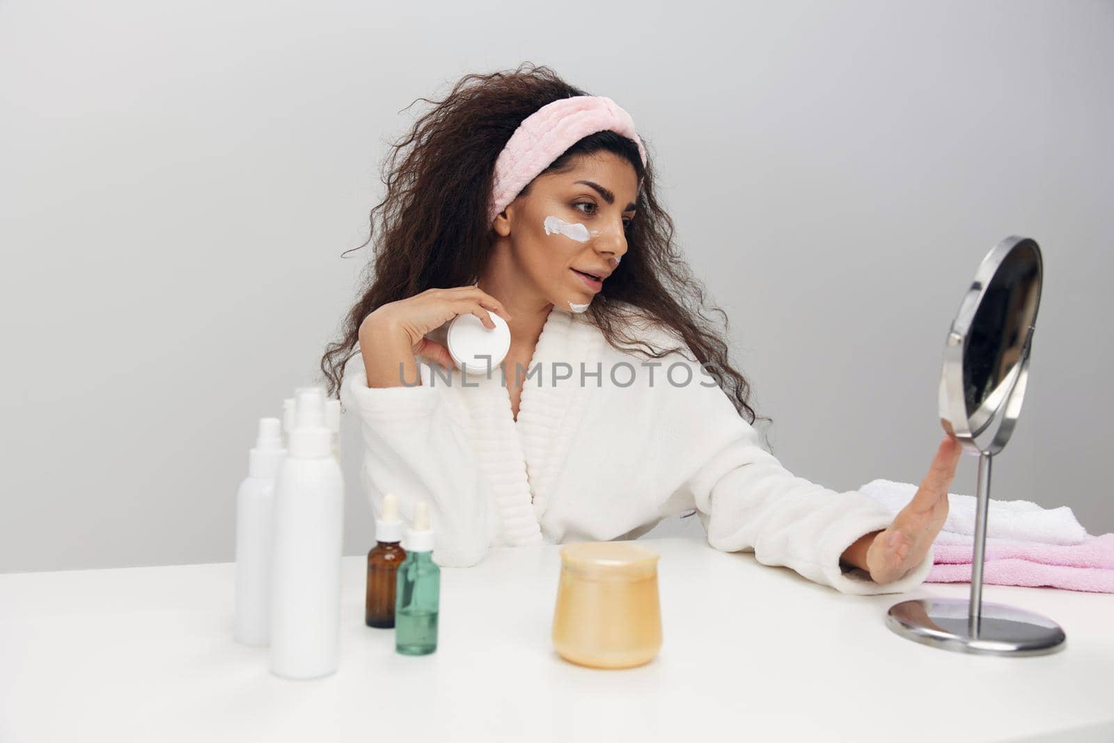 Lovely tanned pretty curly Latin lady admiring herself in mirror use facial cream doing beauty procedure in white home interior. Copy space. MIRROR, MIRROR, ON THE WALL, WHO IS THE FAIREST OF THEM ALL