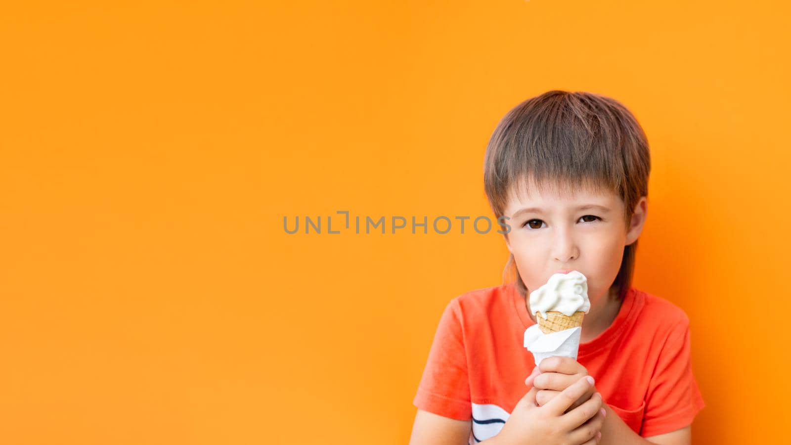 Little boy eats ice cream outdoors. Kid on monochrome, bright orange wall background. Tasty cold dessert at summer.