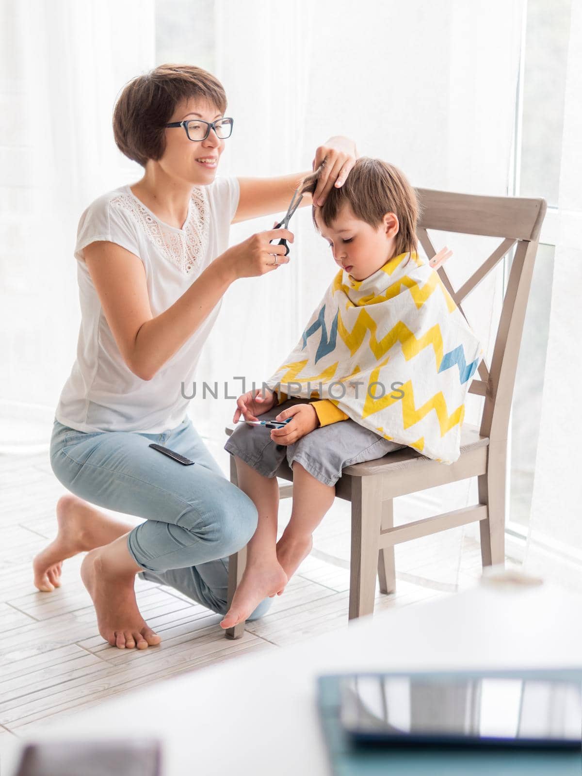 Mother cuts her son's hair by herself. Little boy sits, covered with cloth, and holds pair of scissors. Family life. by aksenovko