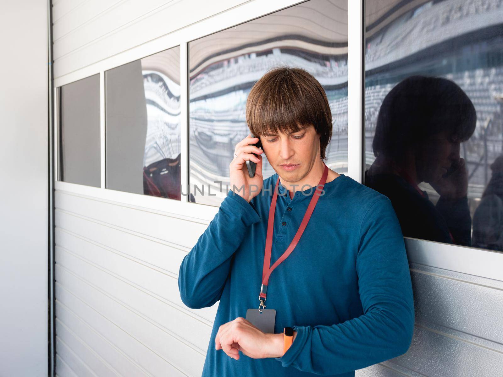 Man with badge on red ribbon talks by smartphone and looks on wristwatch on pit lane at autodrom. Sports car racing. Sports competition on special track.