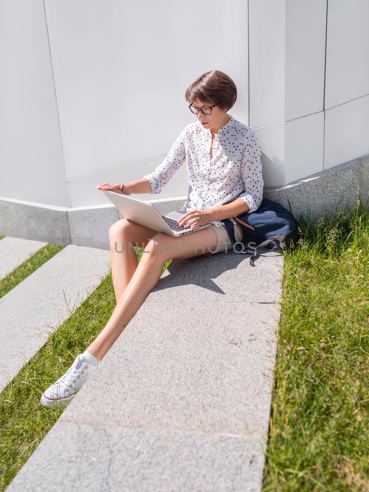 Woman sits with laptop on urban park bench. Freelancer at work. Student learns remotely from outdoors. Modern lifestyle. Summer vibes. Outdoor workplace. by aksenovko