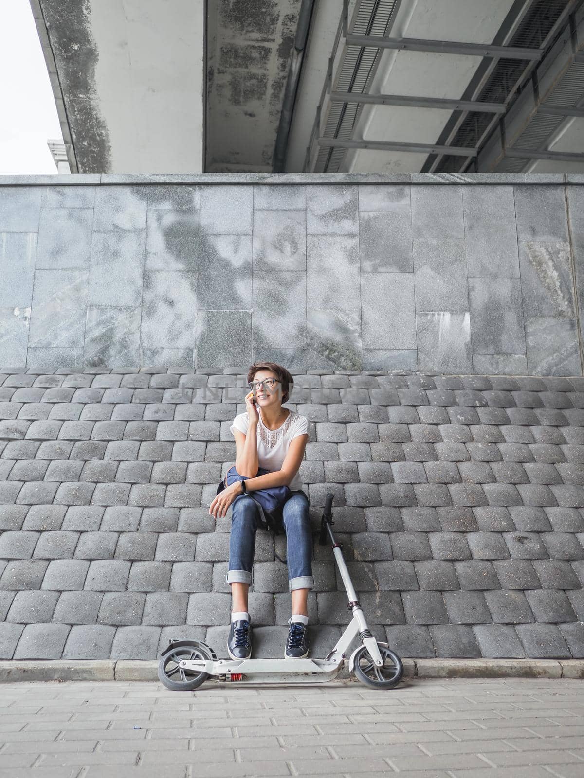 Smiling woman talks by smartphone after riding kick scooter. Female in jeans and sneakers on grey tiled wall background. Healthy lifestyle. Eco-friendly urban transport. by aksenovko