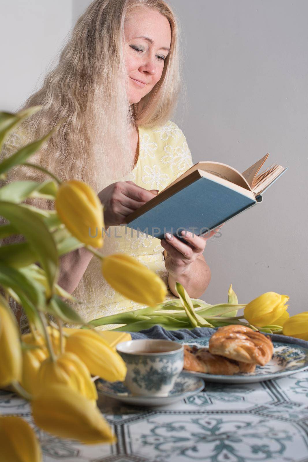 Drinking coffee and tea, a middle-aged blonde woman with long hair drinks tea from a vintage cup and eats puff pastry with pecans on the background of yellow tulips and books on the table