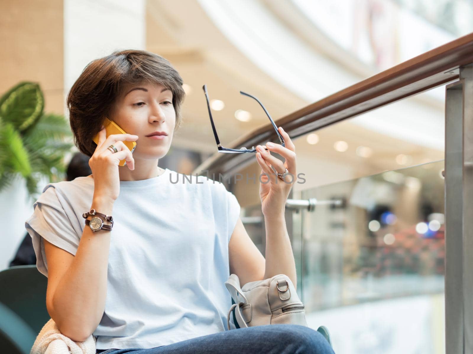Woman is talking by smartphone and is looking surprisingly on her eyeglasses at shopping mall. Coffee break at store.