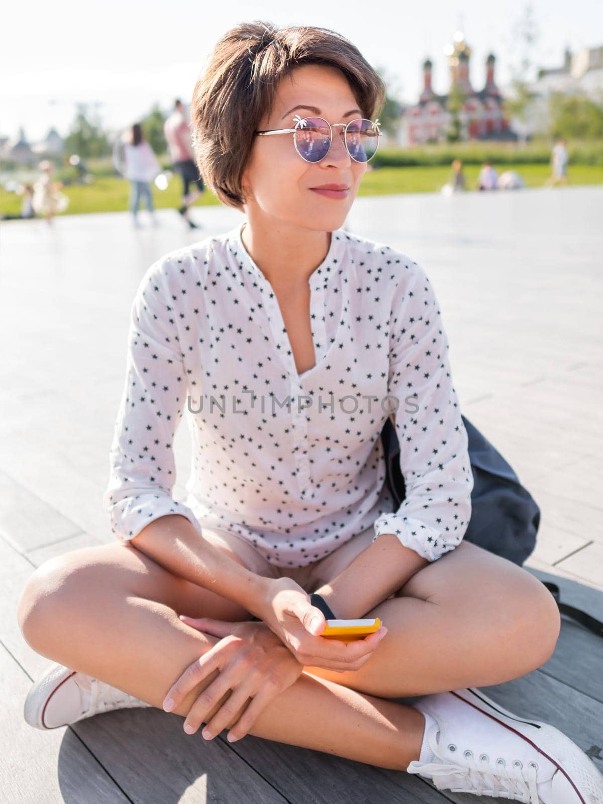 Woman in colorful sunglasses sits on wooden open stage in public park. Pretty female in casual clothes texting on smartphone. by aksenovko