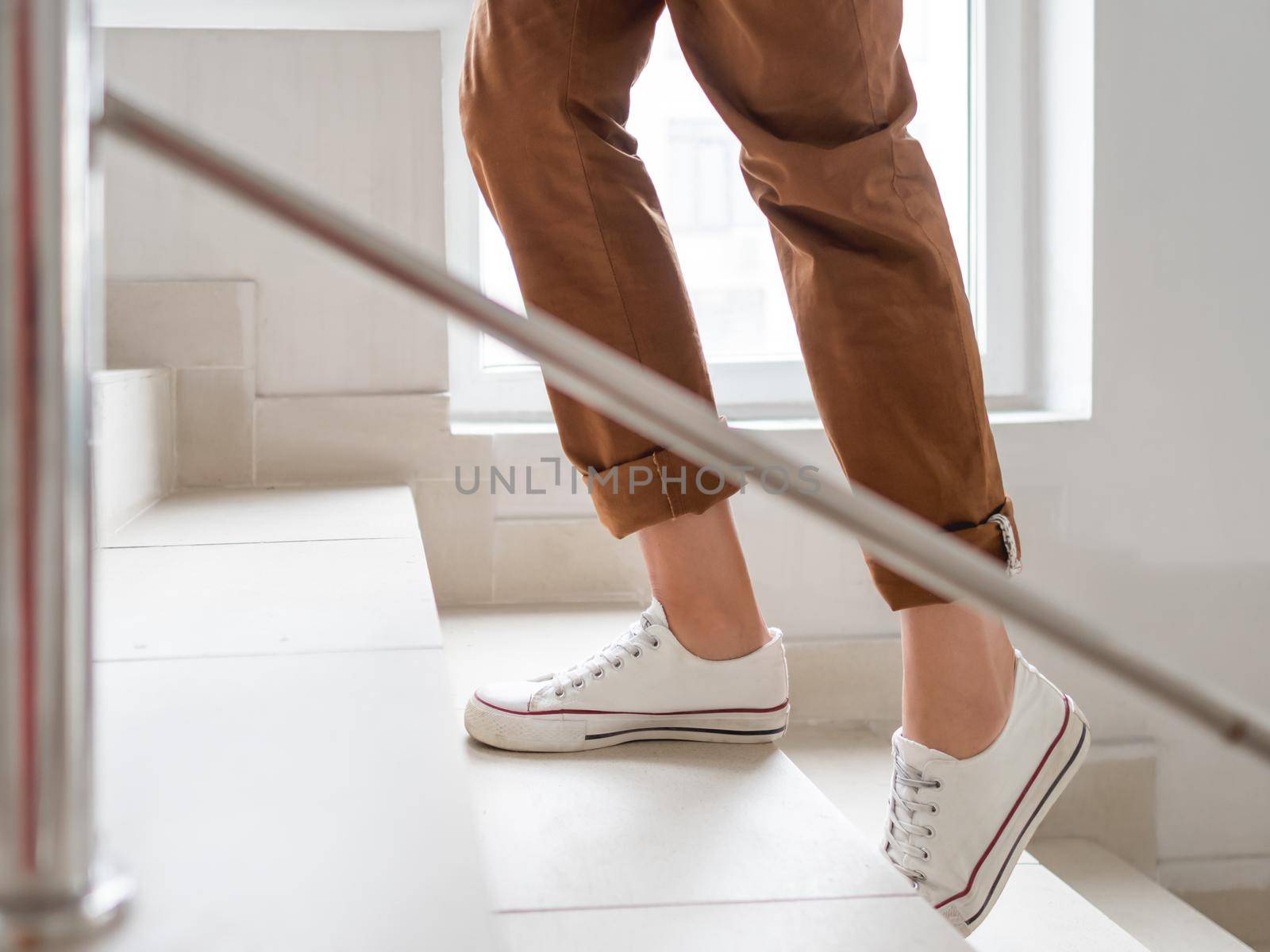 Woman in white sneakers and khaki trousers goes upstairs to her apartment. White staircase in apartment building. Casual outfit, urban fashion. by aksenovko
