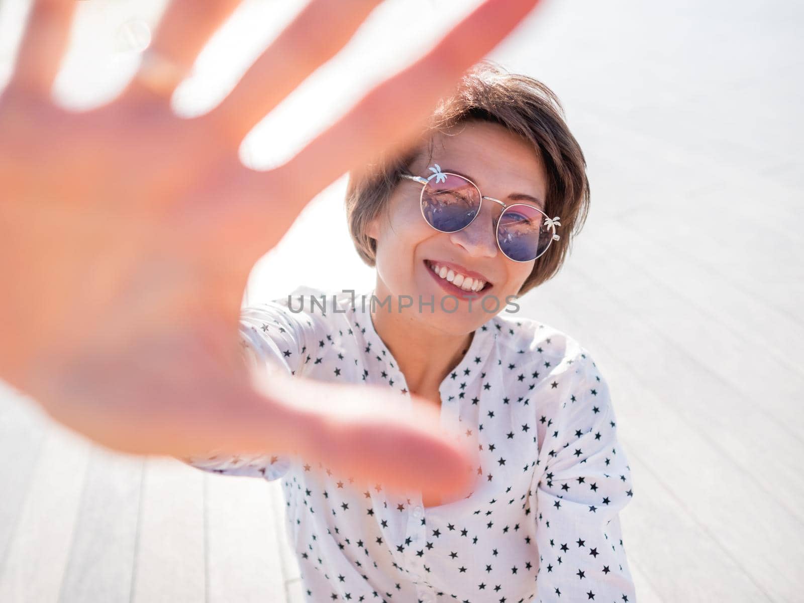 Wind ruffles short hair of freckled woman in colorful sunglasses. Smiling woman at open wooden scene of urban park. Summer vibes. Sincere emotions. by aksenovko