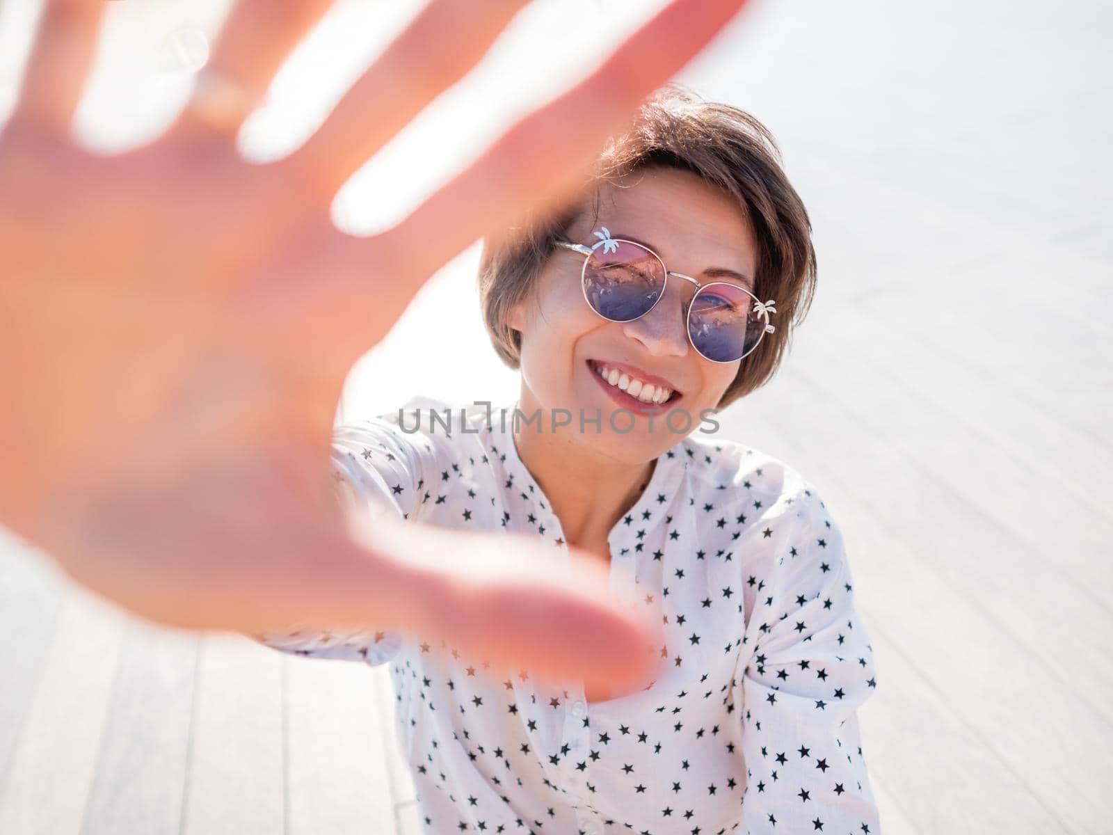 Wind ruffles short hair of freckled woman in colorful sunglasses. Smiling woman at open wooden scene of urban park. Summer vibes. Sincere emotions.