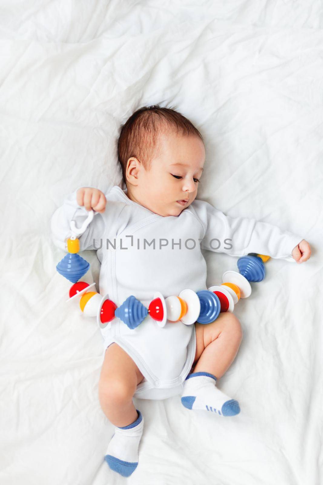 Baby boy holds colorful rattle toy. Top view of little kid lying on crumpled white linen with his first toy. Newborn in bed. by aksenovko