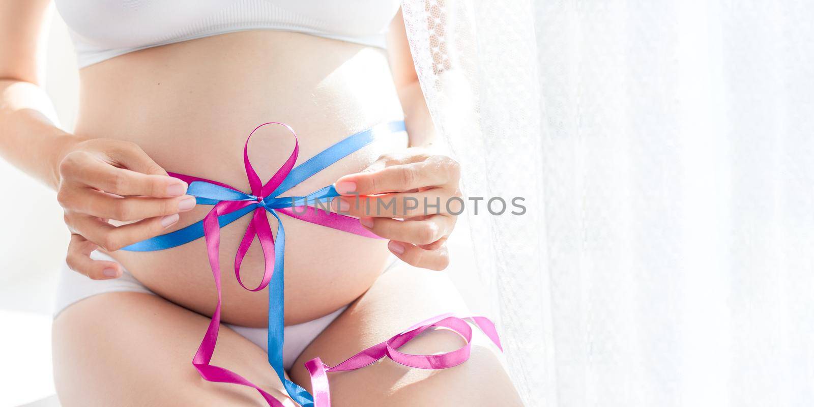 Pregnant woman in white underwear with blue and pink tied bow on belly. Woman in lingerie is expecting twins - boy and girl. Or woman doesn't know sex of future baby.