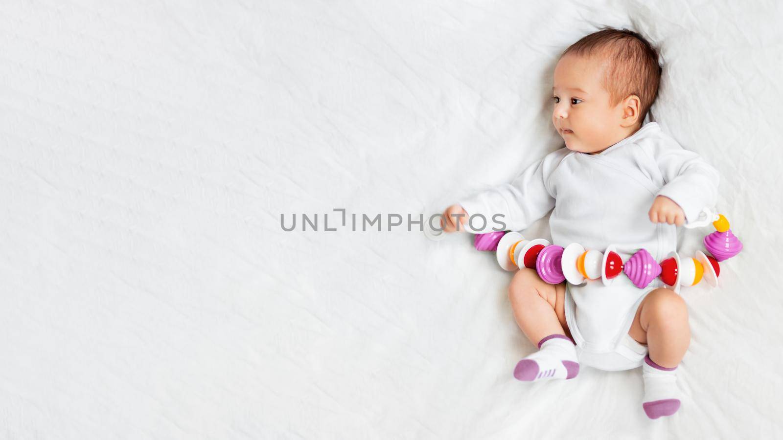 Baby girl holds colorful rattle toy. Top view of little kid lying on crumpled white linen with his first toy. Newborn in bed. Banner with copy space.