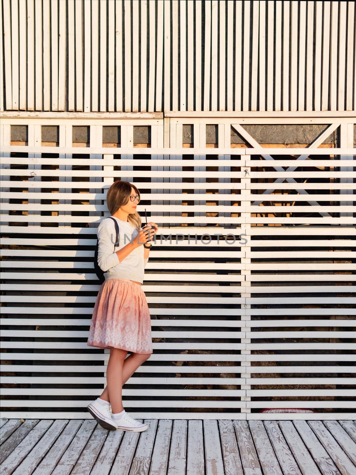 Woman with cup of coffee meets sunset on wooden pier. Female with curly hair and eyeglasses enjoys nature outdoors. Summer casual clothes. Summer vibes.