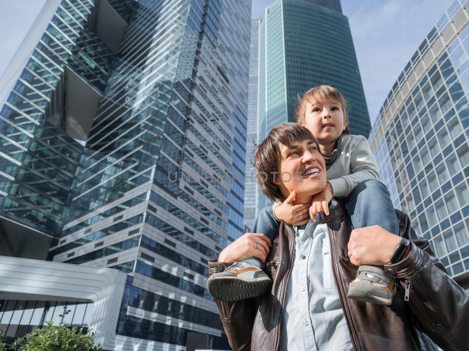 Little boy sits on father's shoulders between skyscrapers. Dad and son looks on glass walls of buildings. Future and modern technologies, life balance and family life in well keeps districts. by aksenovko