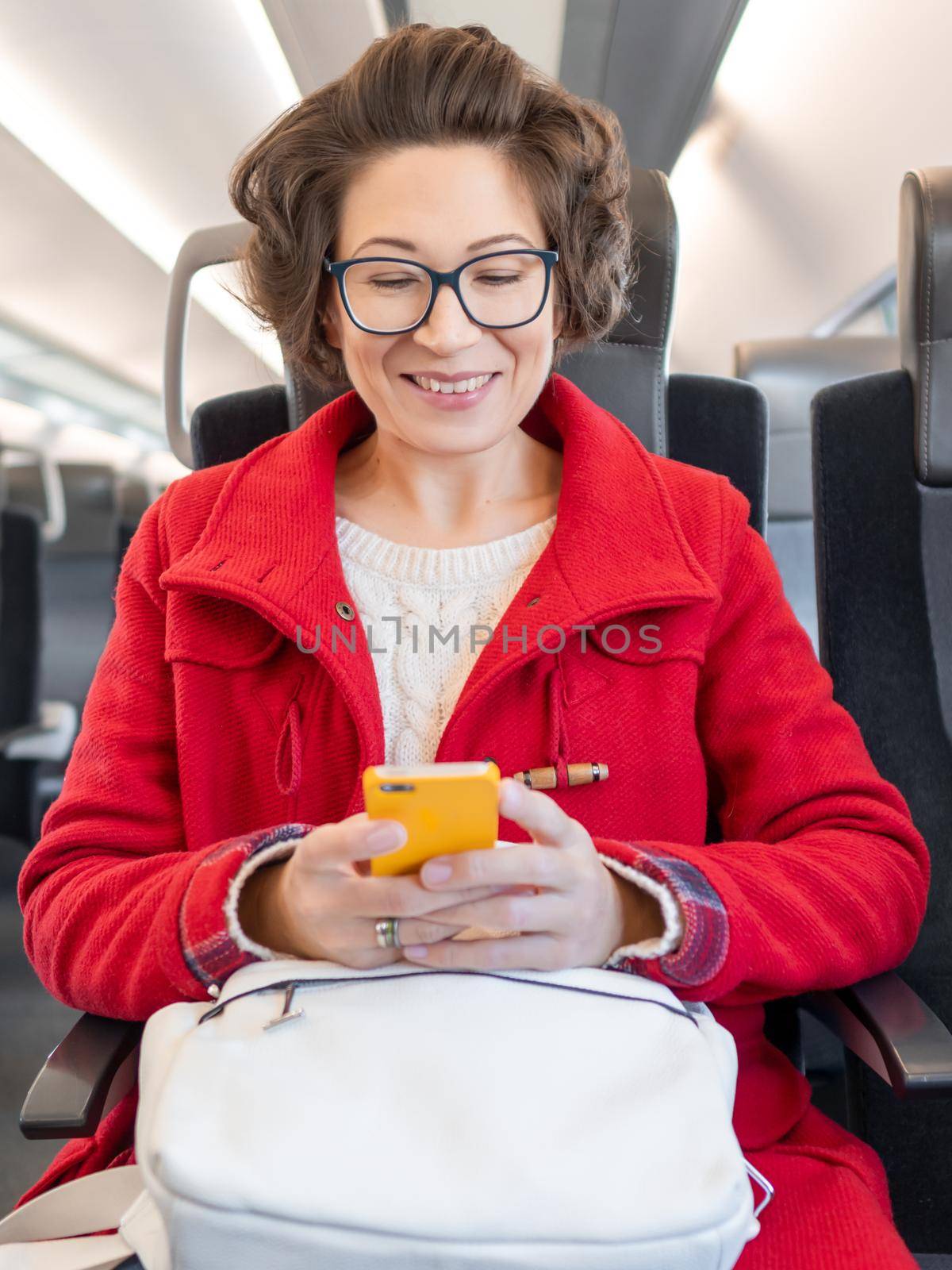 Smiling woman in red duffle coat texting on smartphone in suburban train. Travel by land vehicle.