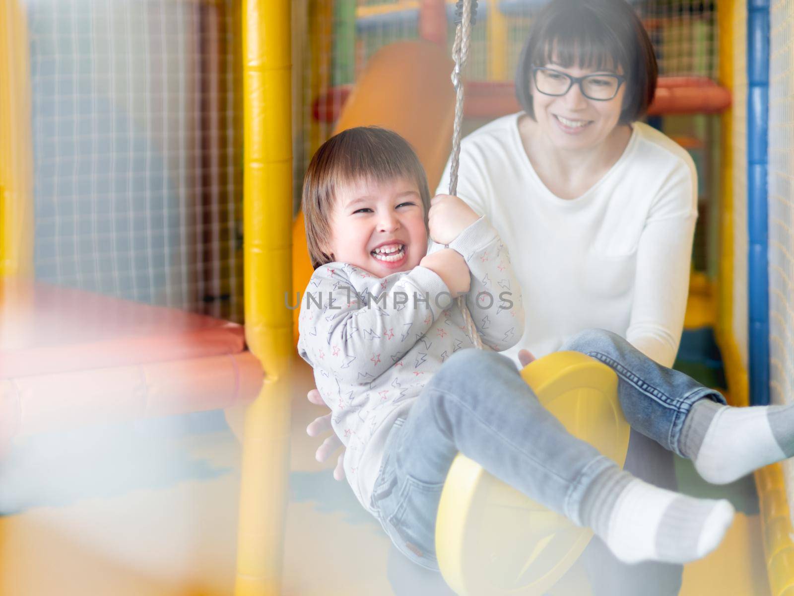 Toddler plays on rope swing with his mother or babysitter. Physical development for little children. Interior of kindergarten or nursery. by aksenovko