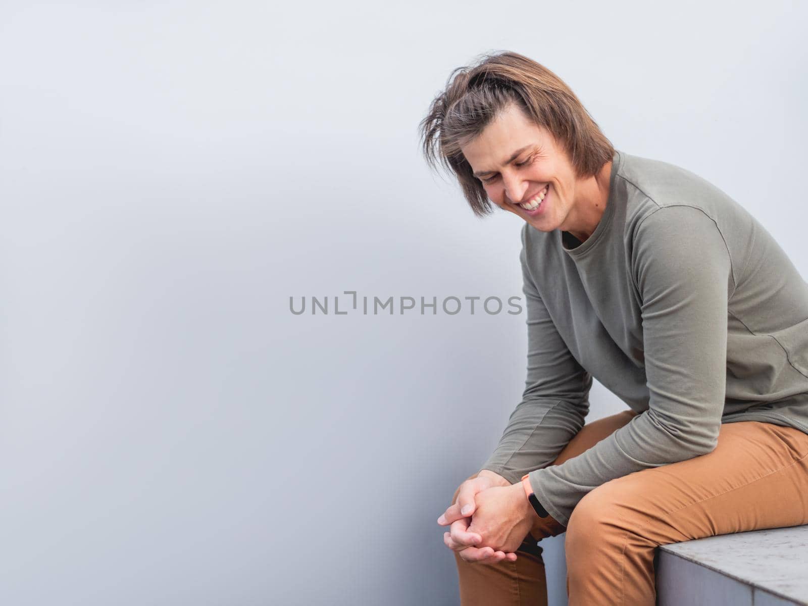 Man with long hair and happy look sits near grey wall. Smiling man in earth toned casual clothes is laughing on horizontal banner with copy space.