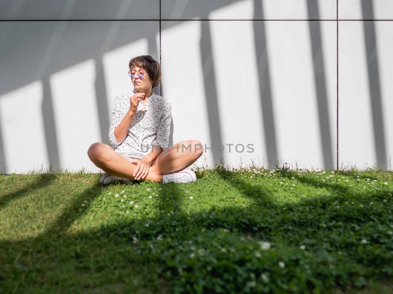 Smiling woman in colorful sunglasses has a rest on lawn in urban park. Nature in town. Relax outdoors after work. Summer vibes.