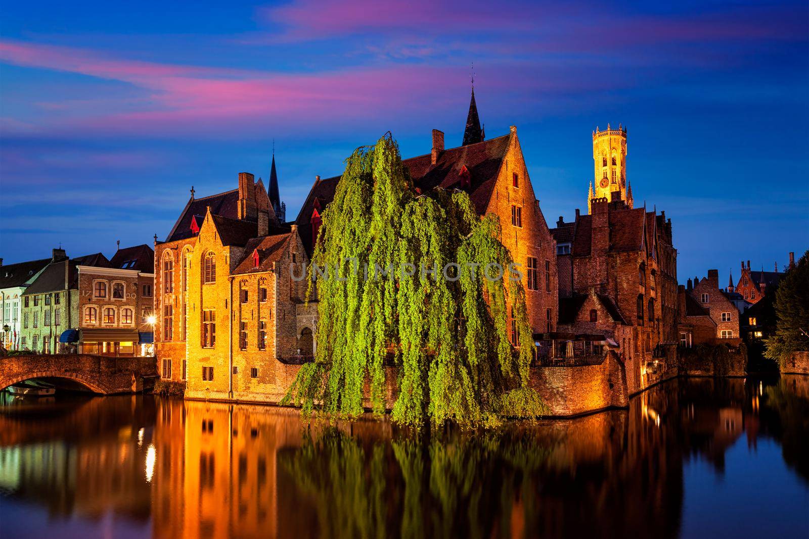 Famous view of Bruges - Rozenhoedkaai with Belfry and old houses along canal with tree in the night. Brugge, Belgium