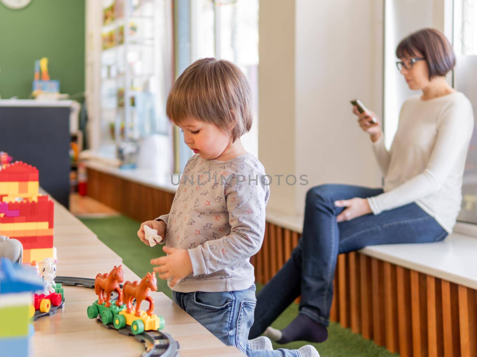 Toddler plays with colorful toy blocks while his mother or babysitter texting in smartphone. Little boy stares on toy constructor. Interior of kindergarten or nursery.