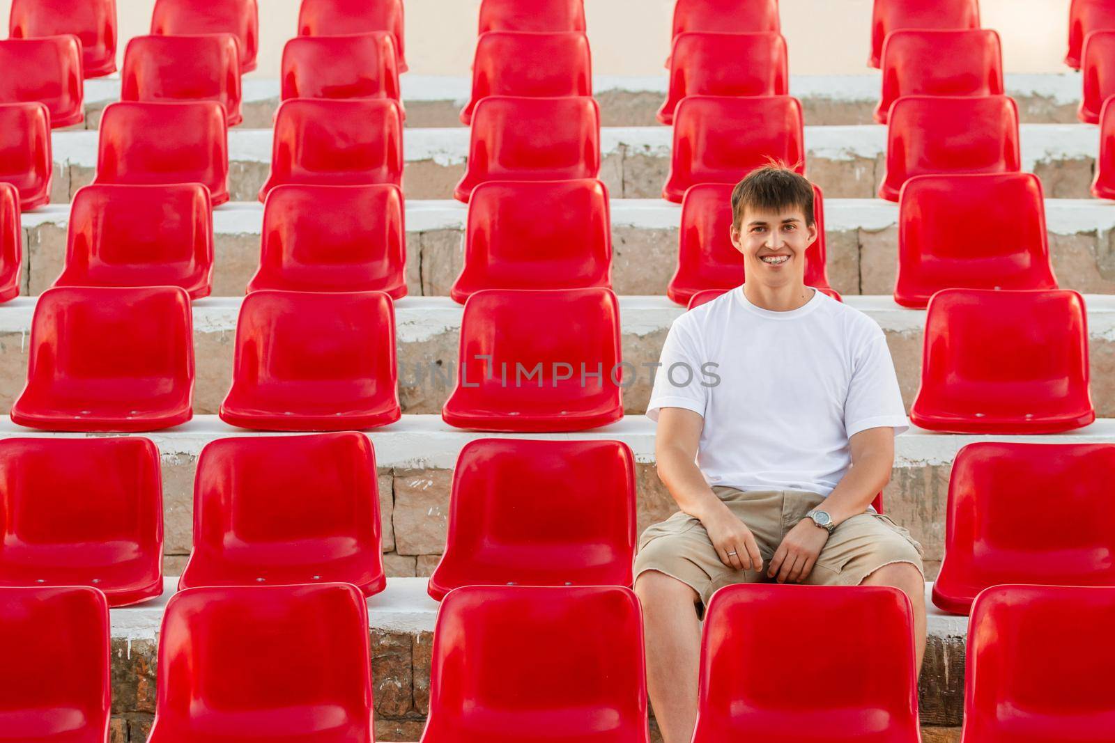 Wide smiling man with dental braces is sitting in deserted audience with bright red seats. Absence of people in open-air amphitheater. by aksenovko