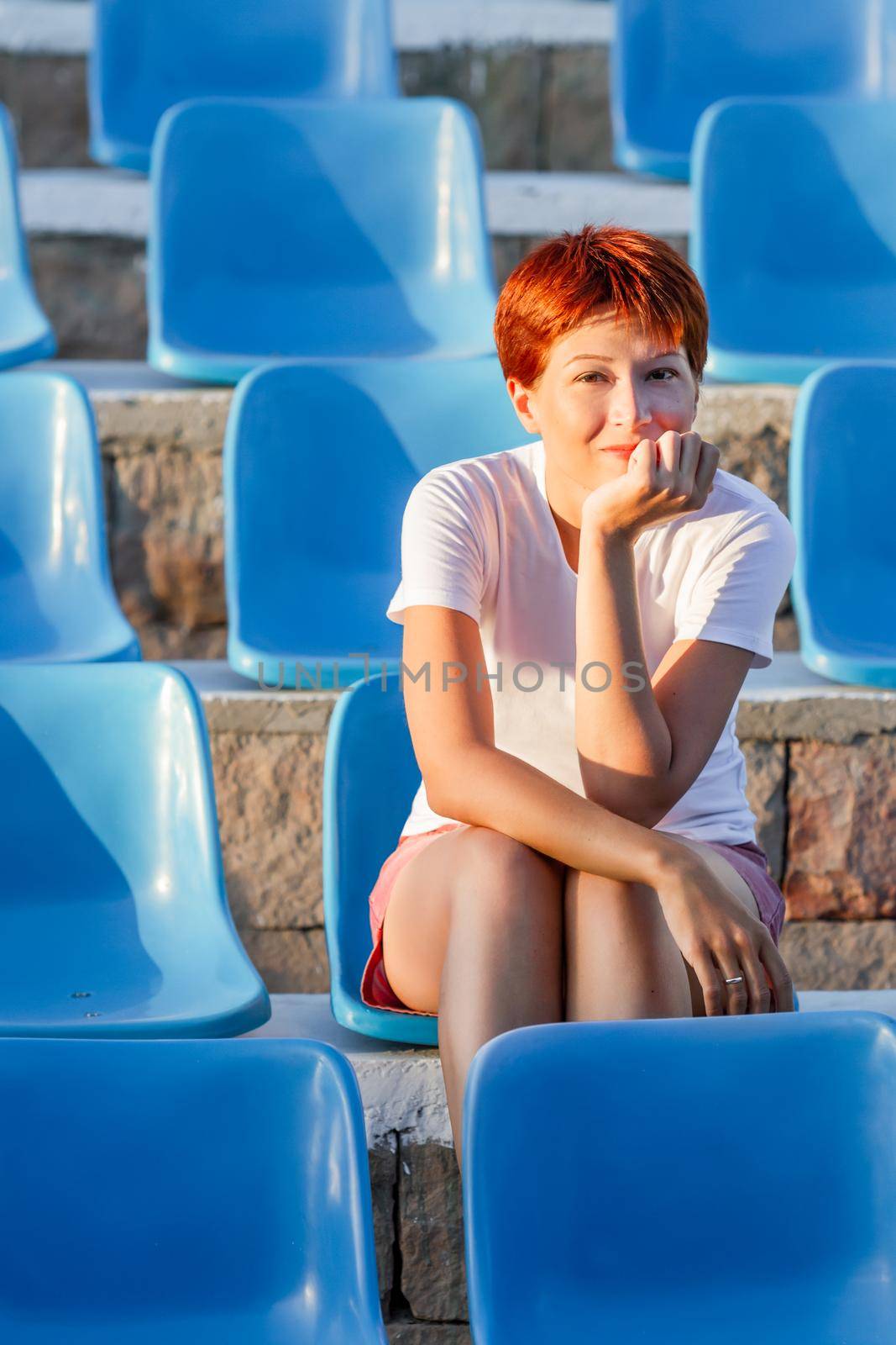 Young woman with short red hair sits relaxed in deserted open air audience. Summer vibes. Sport stadium with plastic blue seats.