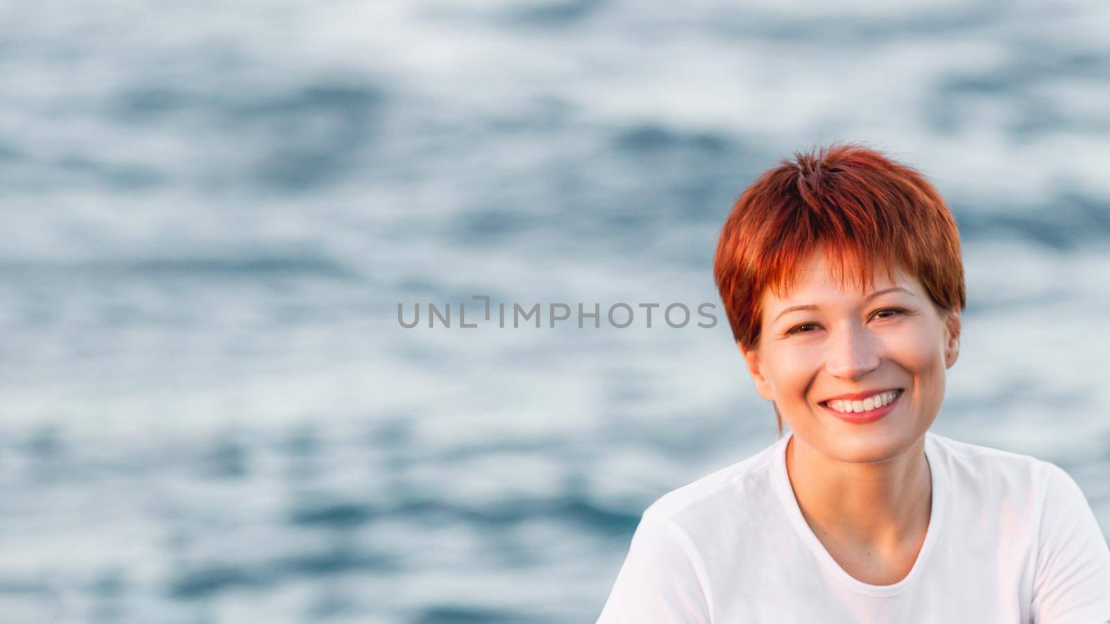 Smiling woman with short bright red haircut. Caucasian woman looks happily on blue sea background. Horizontal banner with copy space.