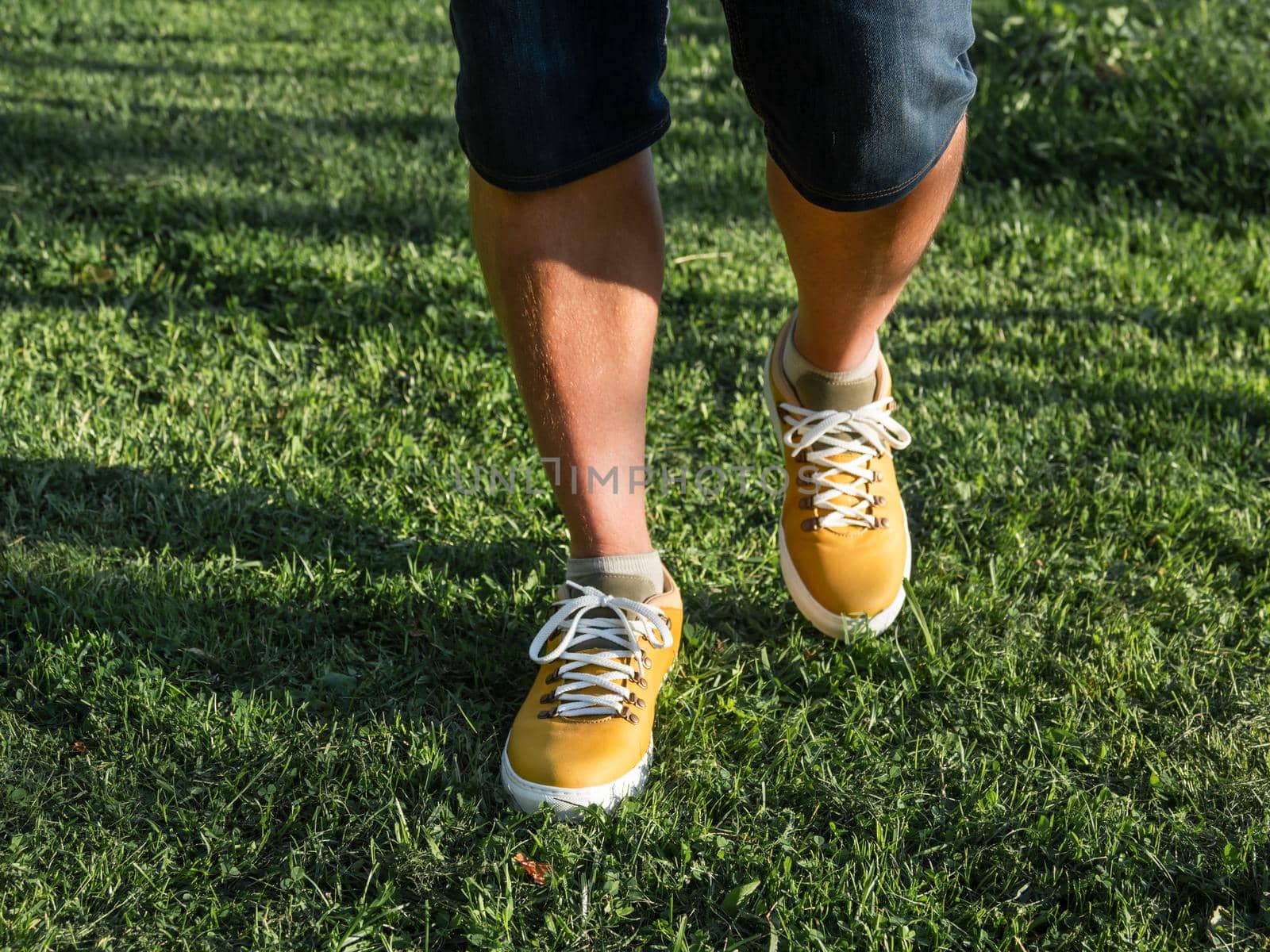Man in bright yellow sneakers is standing on green grass lawn in park. Modern hipster's shoes. Urban fashion.