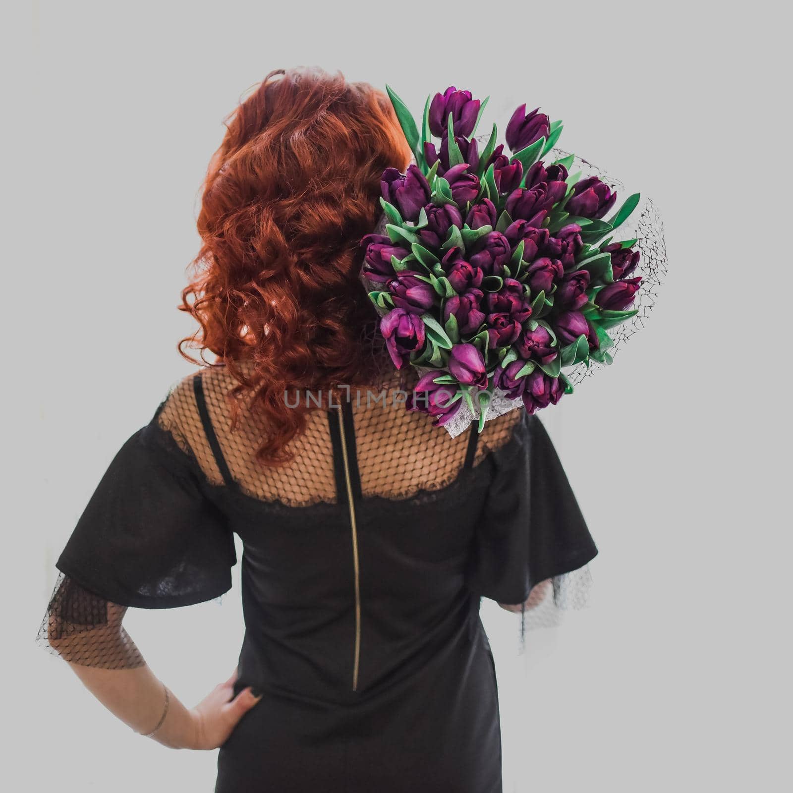 Young beautiful curly-haired red-haired slim girl holds a bouquet of purple tulips on her shoulders while standing backwards. A girl in a black dress with an open back holds her hand at the waist.