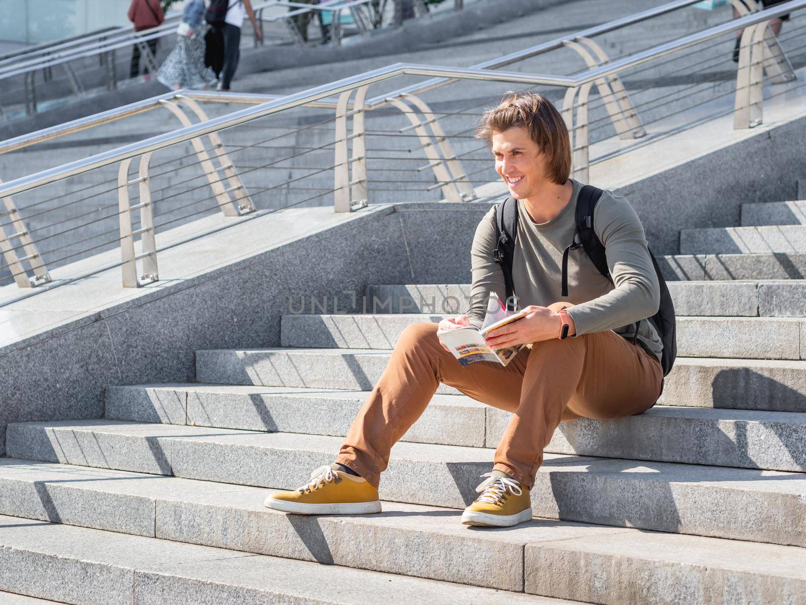 Smiling man sits on stone staircase in park and reads travel guide. Solo-travelling around city. Urban tourism. Modern architectural landmarks. by aksenovko