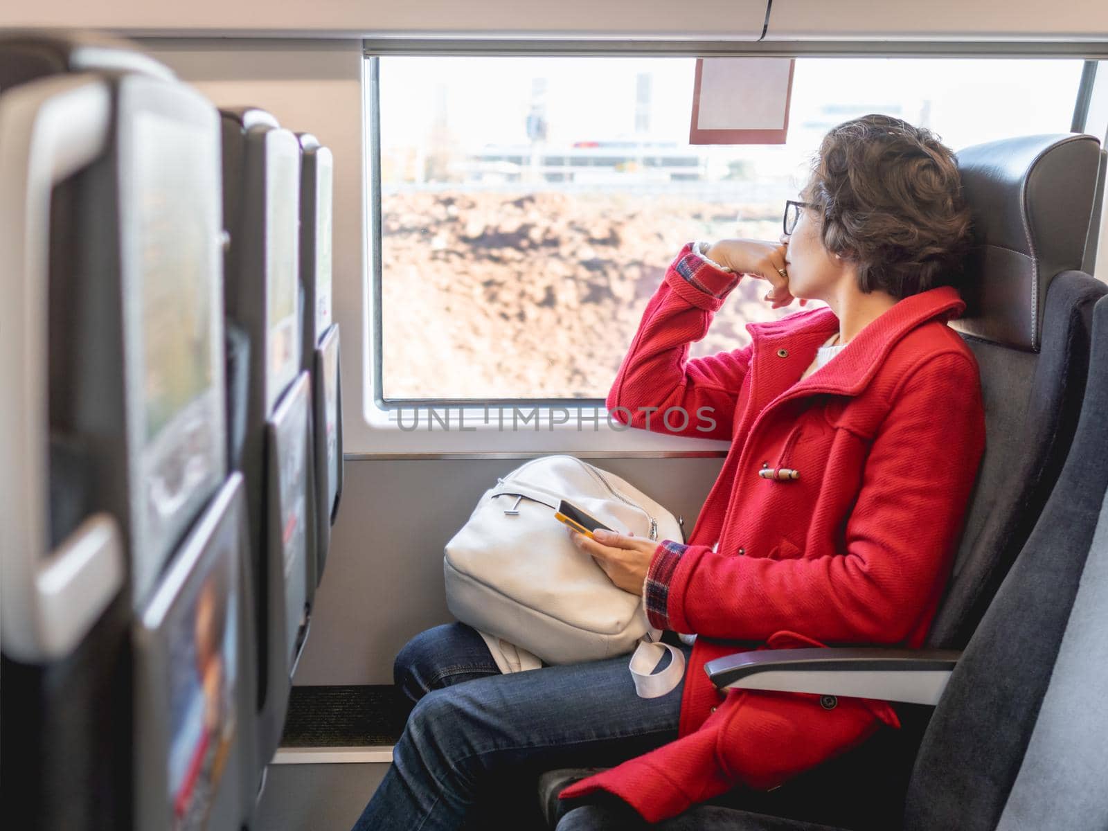 Smiling woman in red duffle coat with on smartphone sits near window in suburban train. Travel by land vehicle. by aksenovko