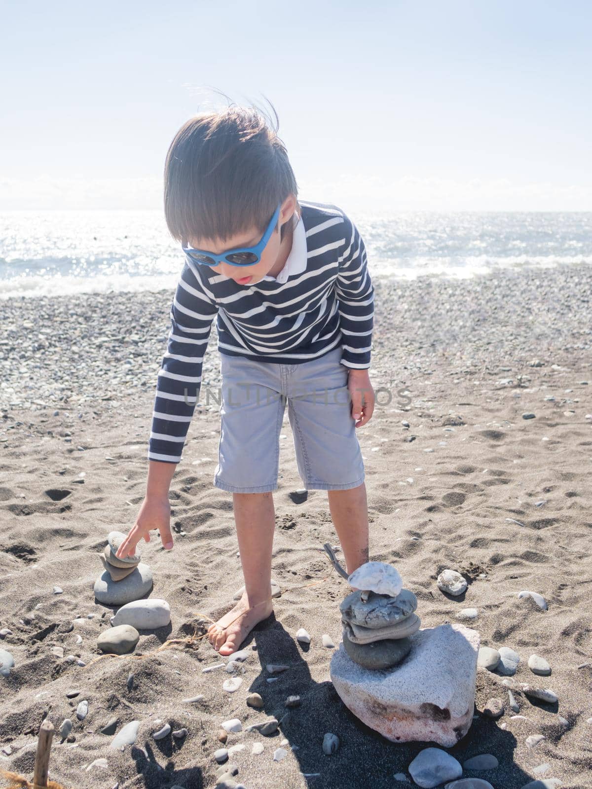 Little boy is building pyramid with stones and driftwood on pebble beach. Vacation on seaside. Exploring nature in childhood.